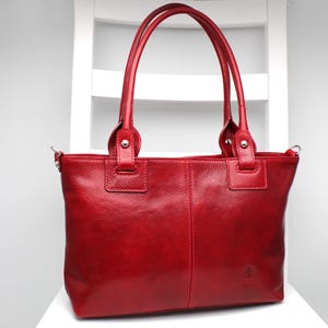 Peut inclure: Un sac cabas en cuir rouge vif avec une surface texturée, deux anses solides fixées par des rivets en métal argenté, une couture verticale centrale et un petit logo en relief sur le coin inférieur droit de la face avant du sac.