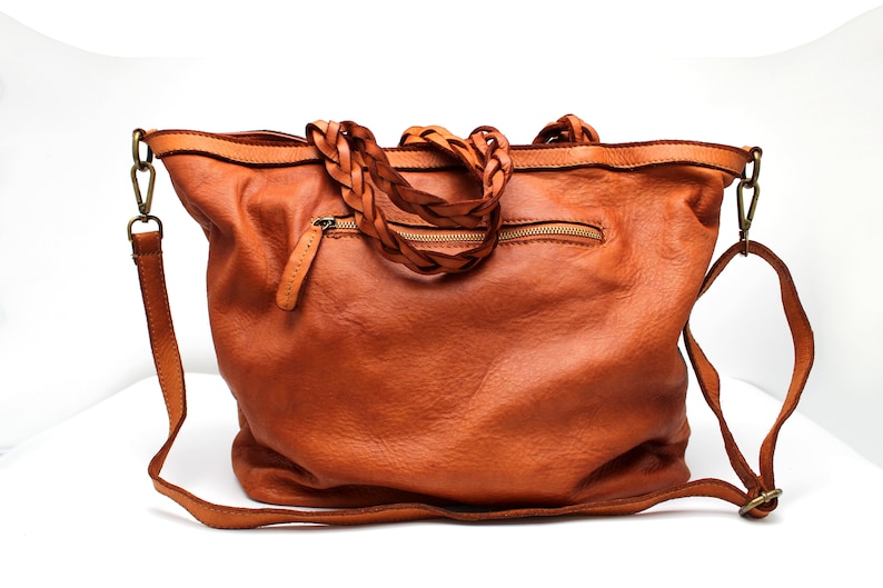 May include: A brown leather tote bag with a braided leather strap. The bag has a zippered pocket on the front and a long shoulder strap.
