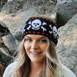 May include: A black knit headband with a white skull and crossbones pattern and orange accents. The headband is worn by a person with long blonde hair.