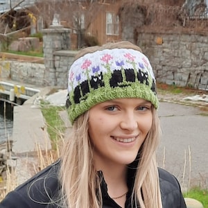 May include: A woman with long blonde hair is wearing a black knitted headband with a floral and cat design. The headband has a green background with white flowers and black cats.