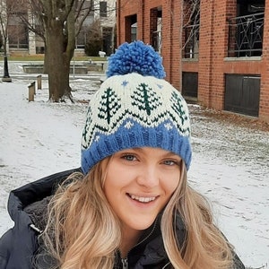 May include: A woman wearing a blue and white knit hat with a green pine tree pattern and a blue pom pom. The hat has a blue and white knit band around the bottom. The woman is smiling and has blonde hair.