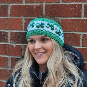 May include: A woman with long blonde hair is wearing a green and white knit headband with a shamrock pattern. She is wearing a black jacket.