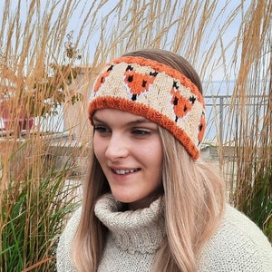 May include: A knitted headband with a pattern of orange fox faces on a cream background. The headband is worn by a person with long blonde hair.