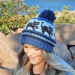 May include: Blue knitted beanie with a pom pom on top and a black cat pattern. The beanie is worn by a person with long blonde hair.