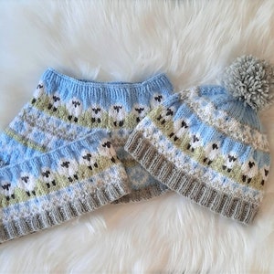 May include: A knitted winter set comprising a light blue hat with a grey pom-pom, a matching neck warmer, and a sweater. The set features a pattern of sheep, white, grey, and green accents. The set is displayed on a white, fluffy surface.
