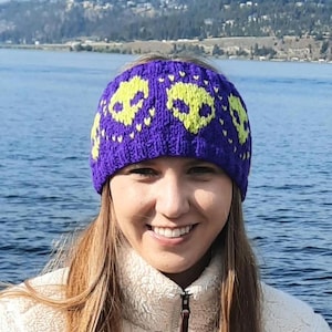 May include: Purple knitted headband featuring a design of yellow alien faces and white hearts. The headband is being worn by a person with long blonde hair.