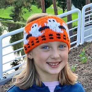 May include: An orange knitted headband featuring white ghost designs with black detailing. The headband is worn by a person with blue eyes and blonde hair. The background includes a lake and trees.