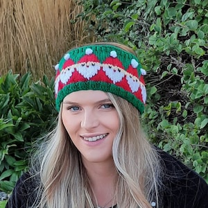 May include: A woman wearing a green and red knit headband with a pattern of Santa faces. The headband has white pom-poms on top.