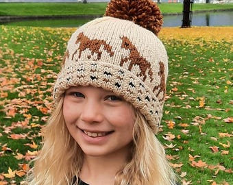 May include: A beige knit hat with brown horse silhouettes and a large brown pom-pom. The hat features a patterned brim and is worn by a person with blonde hair, set against a backdrop of autumn foliage and a lake.