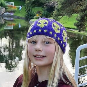 May include: A person wearing a purple knitted hat featuring a repeating pattern of yellow skulls. The hat is worn with a burgundy zip-up jacket. The background shows a lake, trees, and a park setting.