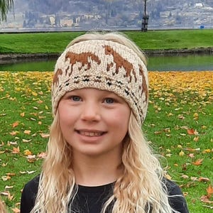 Peut inclure: Bandeau tricoté beige et marron orné d'un motif de chevaux répété. Porté par une personne aux cheveux blonds, sur fond d'herbe verte, d'eau et d'arbres, évoquant une scène automnale.