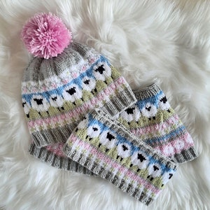 May include: A gray knitted hat and matching scarf with a pink pom-pom on top. The hat and scarf feature a white sheep pattern with black eyes and a pink, blue, and green color scheme.