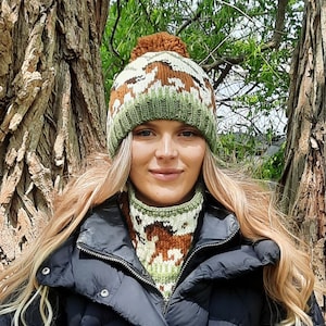 May include: A woman wearing a brown and green knitted hat with a pom pom and a matching knitted neck warmer. The hat and neck warmer feature a repeating pattern of deer.