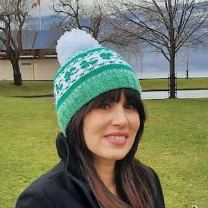 May include: A green and white knit hat with a white pom pom on top. The hat has a pattern of green and white squares. The hat is being worn by a person with long brown hair.