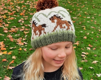May include: A cream-colored knit hat with a brown pom-pom and a green brim. The hat features a repeating pattern of brown horses and green accents. The hat is worn by a person with blonde hair.