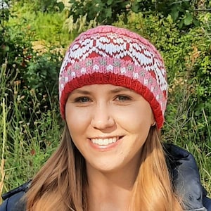 May include: A woman wearing a pink and white knitted hat with a geometric pattern. The hat has a red band around the bottom.