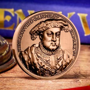 Könnte beinhalten: Eine bronzefarbene Münze mit einem detaillierten Porträt einer historischen Figur, möglicherweise eines Königs, mit aufwendiger Kleidung und Hut. Die Münze hat Inschriften am Rand. Im Hintergrund ein blaues Buch mit goldfarbenen Buchstaben.