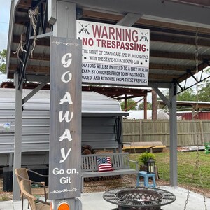 Op de afbeelding: Een houten bord met de tekst "WARNING NO TRESPASSING." Het bord is bevestigd aan een houten paal met een tweede bord waarop staat "GO AWAY Go on, Git". De borden staan voor een overdekt terras met een vuurplaats, stoelen en een schommel.