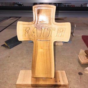Puede incluir: Una cruz de madera con la palabra "FAITH" tallada en ella. La cruz está de pie sobre una base de madera.