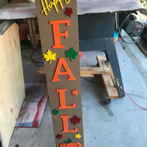 May include: A wooden sign with the words "Happy Fall" in orange letters. The sign is decorated with fall leaves in shades of orange, yellow, and green. A pumpkin is at the bottom of the sign.