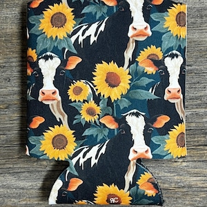 May include: A black and white cow-themed beverage holder with a sunflower pattern. The design features multiple cow faces and yellow sunflowers against a dark blue background. The bottom of the holder has a curved cutout.