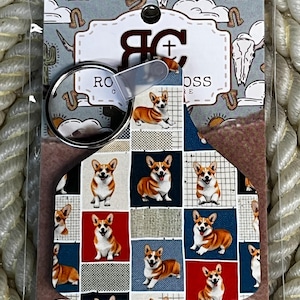 May include: A keychain featuring a tag with a repeating pattern of Corgi dogs on a background of red, blue, and white grid squares. The tag is attached to a metal ring and mounted on a display card labeled Rock Cross with western-themed graphics.