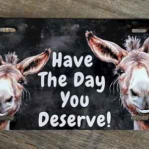May include: Rectangular license plate featuring two smiling donkeys with large ears. The plate has a black background with the text "Have The Day You Deserve!" in white. The donkeys are painted in shades of brown and gray.