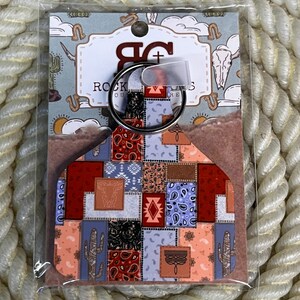May include: A keychain shaped like a livestock ear tag featuring a colorful patchwork design of paisley, geometric patterns, and cactus illustrations. The tag is attached to a silver metal ring and mounted on a branded card with the text ROCK + SOUL.