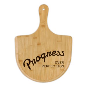 May include: A bamboo cutting board with a handle and a curved bottom. The board has the words "Progress Over Perfection" in black script. The board is made of light brown bamboo.