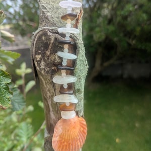May include: A vertical hanging ornament featuring a series of smooth, frosted sea glass pieces in shades of white, light blue, and dark brown, interspersed with small clear beads, ending with a large, textured, fan-shaped orange seashell.