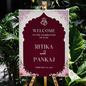 May include: A welcome sign for a wedding with a burgundy background and white text. The sign says "Welcome to the celebration of love Ritika and Pankaj February 25, 2025". The sign is decorated with pink flowers and a traditional Indian design.