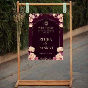 May include: A wedding welcome sign with a burgundy background and gold accents. The sign features floral designs and the text "Welcome to the celebration of love Ritika and Pankaj February 25, 2025".