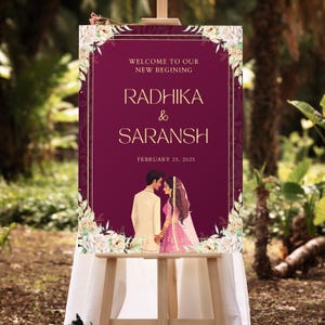 May include: A wedding welcome sign with a burgundy background and gold accents. The sign features a floral design with white flowers and green leaves. The text on the sign reads "Welcome to our new beginning, Radhika & Saransh, February 25, 2025".