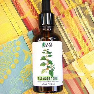 Może przedstawiać: Brązowa szklana butelka z czarną pipetą, z etykietą "Lucky Forest" i "Ashwagandha Double Extraction Tincture". Etykieta zawiera ilustrację botaniczną. Butelka zawiera 50 ml płynu. Tło to kolorowa patchworkowa tkanina.