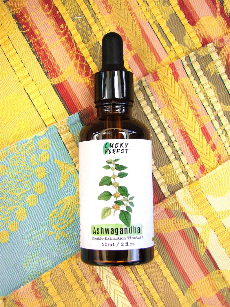 Może przedstawiać: Brązowa szklana butelka z czarną pipetą, z etykietą "Lucky Forest" i "Ashwagandha Double Extraction Tincture". Etykieta zawiera ilustrację botaniczną. Butelka zawiera 50 ml płynu. Tło to kolorowa patchworkowa tkanina.