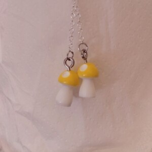 May include: A pair of dangle earrings featuring mushroom charms. Each charm has a yellow cap with white spots and a white stem. The earrings hang from silver-colored chains.