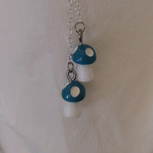 May include: A silver chain necklace featuring two mushroom pendants. The mushroom caps are teal with white spots, and the stems are white. The pendants are connected to the chain with silver jump rings.