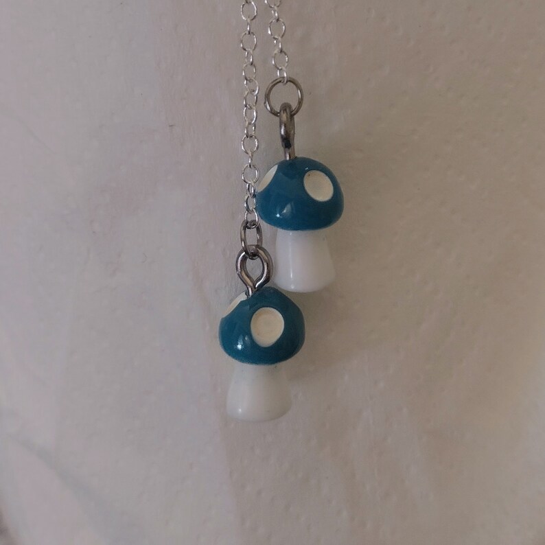 May include: A silver chain necklace featuring two mushroom pendants. The mushroom caps are teal with white spots, and the stems are white. The pendants are connected to the chain with silver jump rings.
