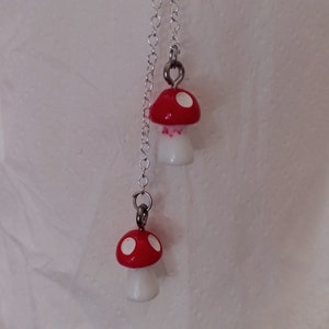 May include: A pair of mushroom-shaped earrings. Each earring features a red cap with white spots and a white stem. The earrings are connected by a silver chain and are designed to dangle. The background is a plain white surface.