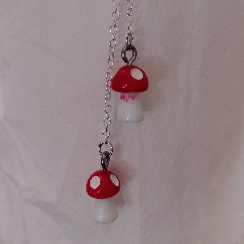 May include: A pair of mushroom-shaped earrings. Each earring features a red cap with white spots and a white stem. The earrings are connected by a silver chain and are designed to dangle. The background is a plain white surface.