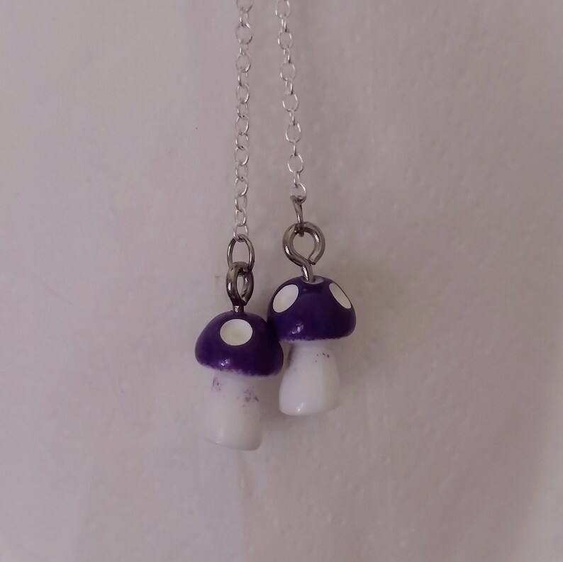 May include: A pair of dangle earrings featuring miniature mushroom charms. The mushroom caps are purple with white spots, and the stems are white. The earrings hang from delicate silver chains.