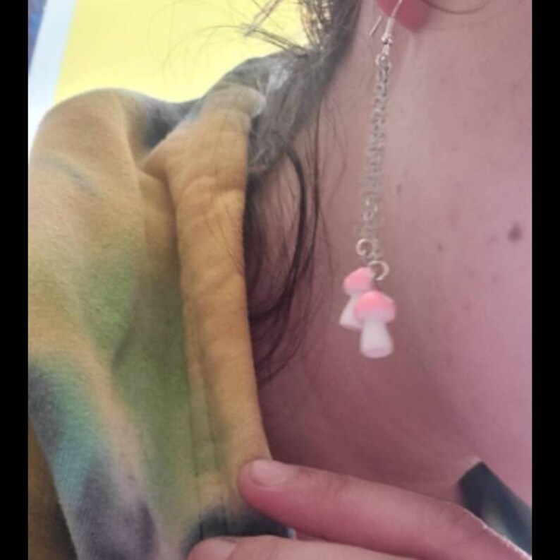 May include: A silver chain earring with two small mushroom charms. The mushroom caps are pink, and the stems are white. The earring is hanging from a silver hook.