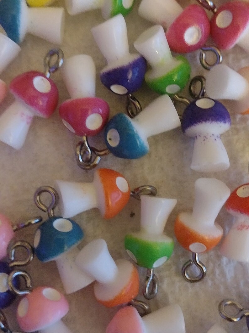 May include: A collection of colorful mushroom charms with white stems and caps in various colors, including pink, blue, green, and orange. Each charm has a silver-colored loop for attaching to jewelry or other crafts. The mushroom caps have white polka dots.