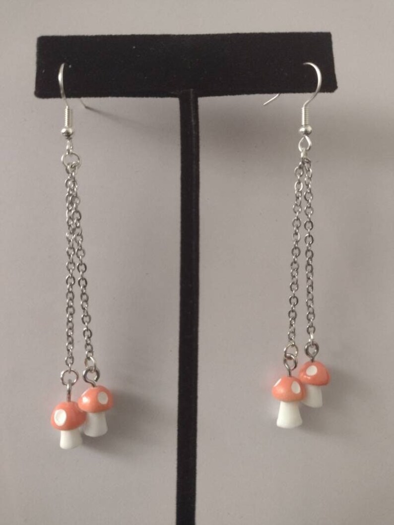 May include: A pair of dangle earrings featuring small, orange mushroom charms with white spots and stems. The mushroom charms are suspended from silver-colored chain and hooks. The earrings are displayed on a black stand against a neutral background.
