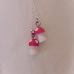 May include: A silver chain necklace featuring two mushroom pendants. The mushroom caps are bright red with white spots, and the stems are white. The necklace is a whimsical accessory.