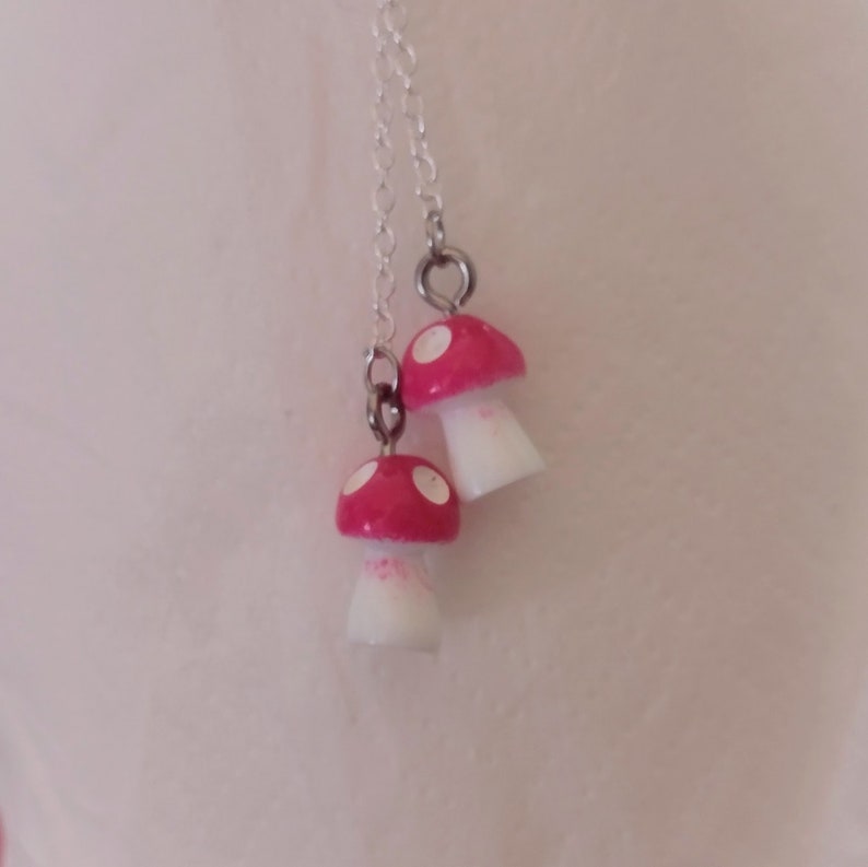 May include: A silver chain necklace featuring two mushroom pendants. The mushroom caps are bright red with white spots, and the stems are white. The necklace is a whimsical accessory.