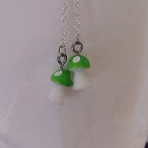 May include: A pair of dangle earrings featuring mushroom charms. The mushroom caps are bright green with white spots, and the stems are white. The earrings hang from delicate silver chains.