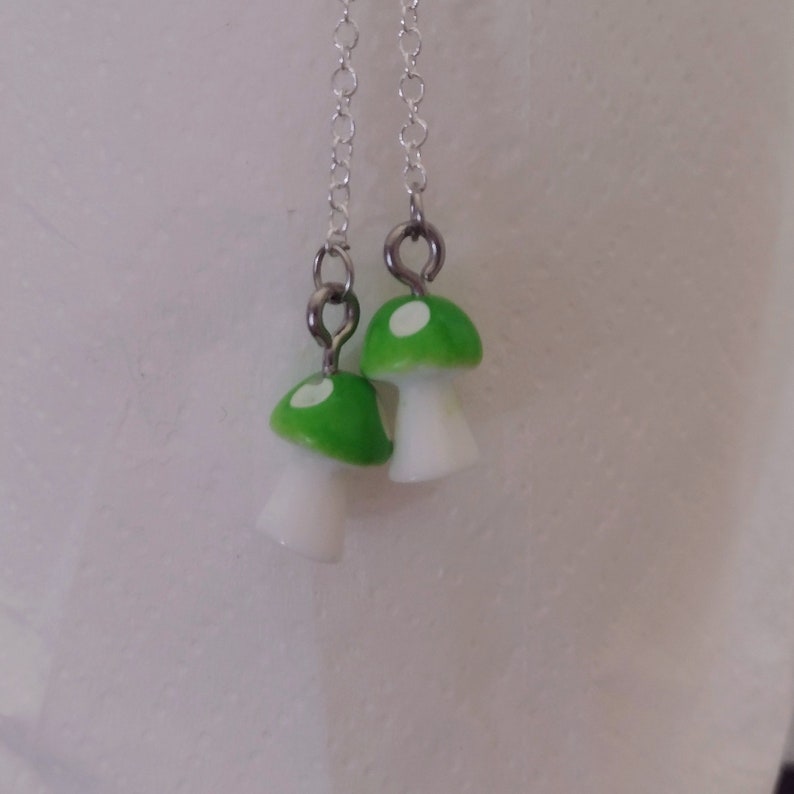 May include: A pair of dangle earrings featuring mushroom charms. The mushroom caps are bright green with white spots, and the stems are white. The earrings hang from delicate silver chains.