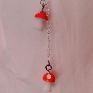 May include: A silver chain necklace featuring two mushroom pendants. The mushroom caps are bright red with white spots, and the stems are white. The necklace is suspended against a soft, off-white background.