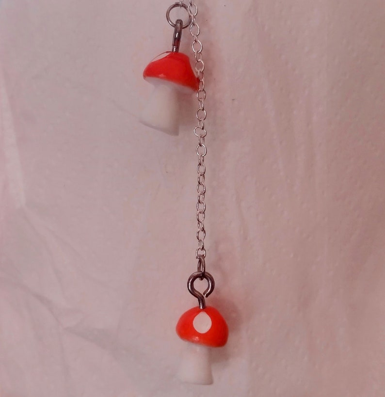 May include: A silver chain necklace featuring two mushroom pendants. The mushroom caps are bright red with white spots, and the stems are white. The necklace is suspended against a soft, off-white background.